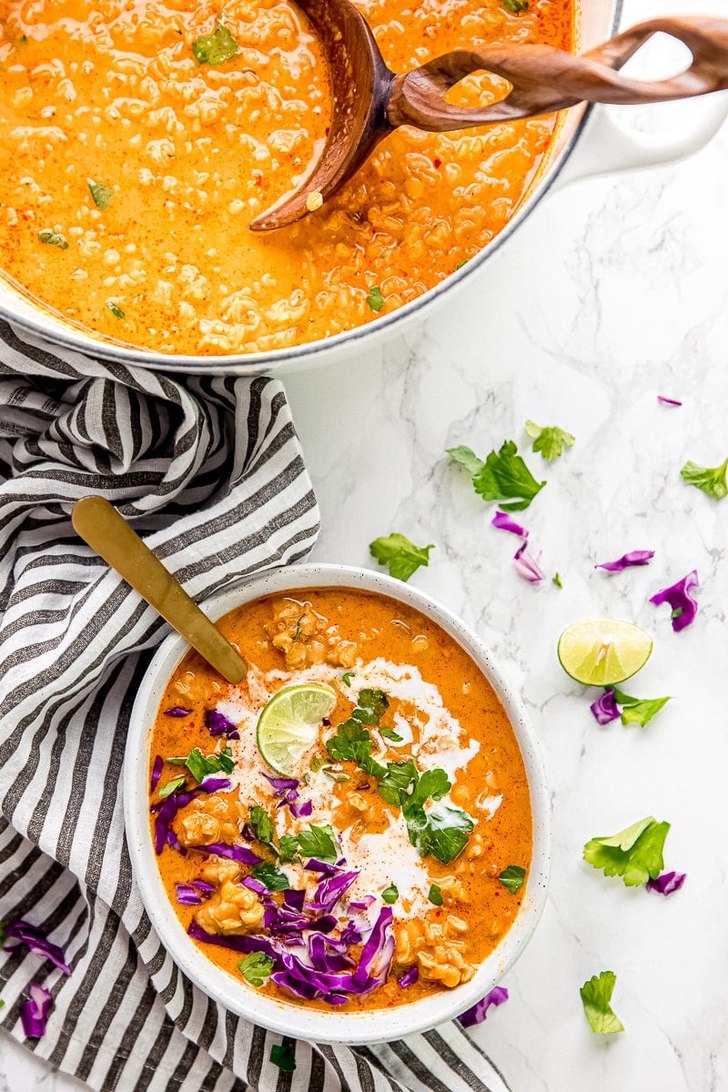 A bowl of coconut curry lentil soup topped with parsley, coconut milk, cabbage, and lime, with a spoon in it, next to a serving bowl of soup with a serving spoon