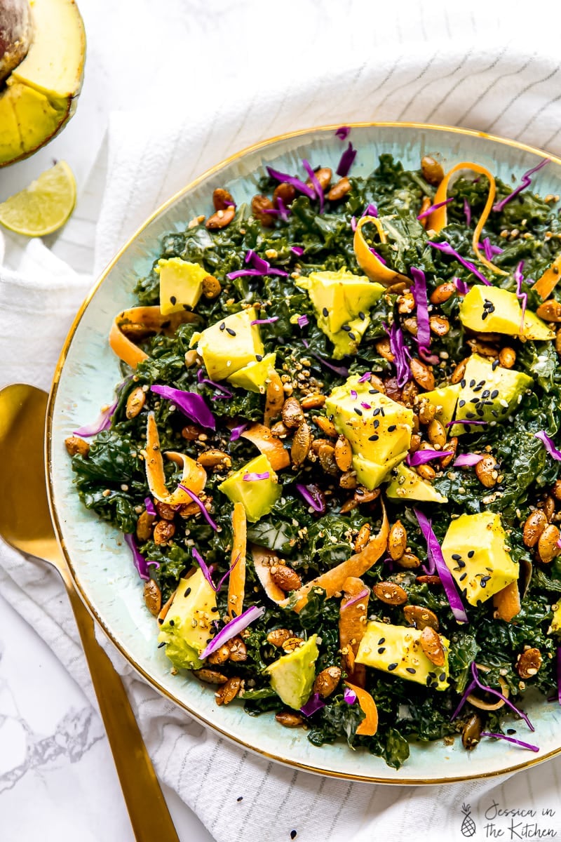 Overhead view of kale salad topped with avocado, carrots, seeds, and dressing in a large serving bowl.