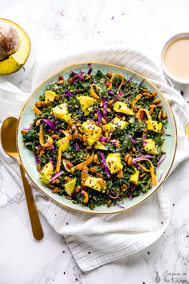 Overhead view of kale salad topped with avocado, carrots, seeds, and dressing in a large serving bowl, next to a spoon and a small bowl of tahini dressing.