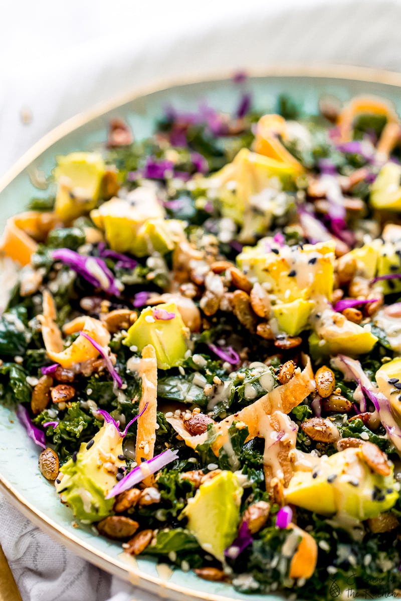 Close up of kale salad topped with avocado, carrots, seeds, and dressing in a large serving bowl.