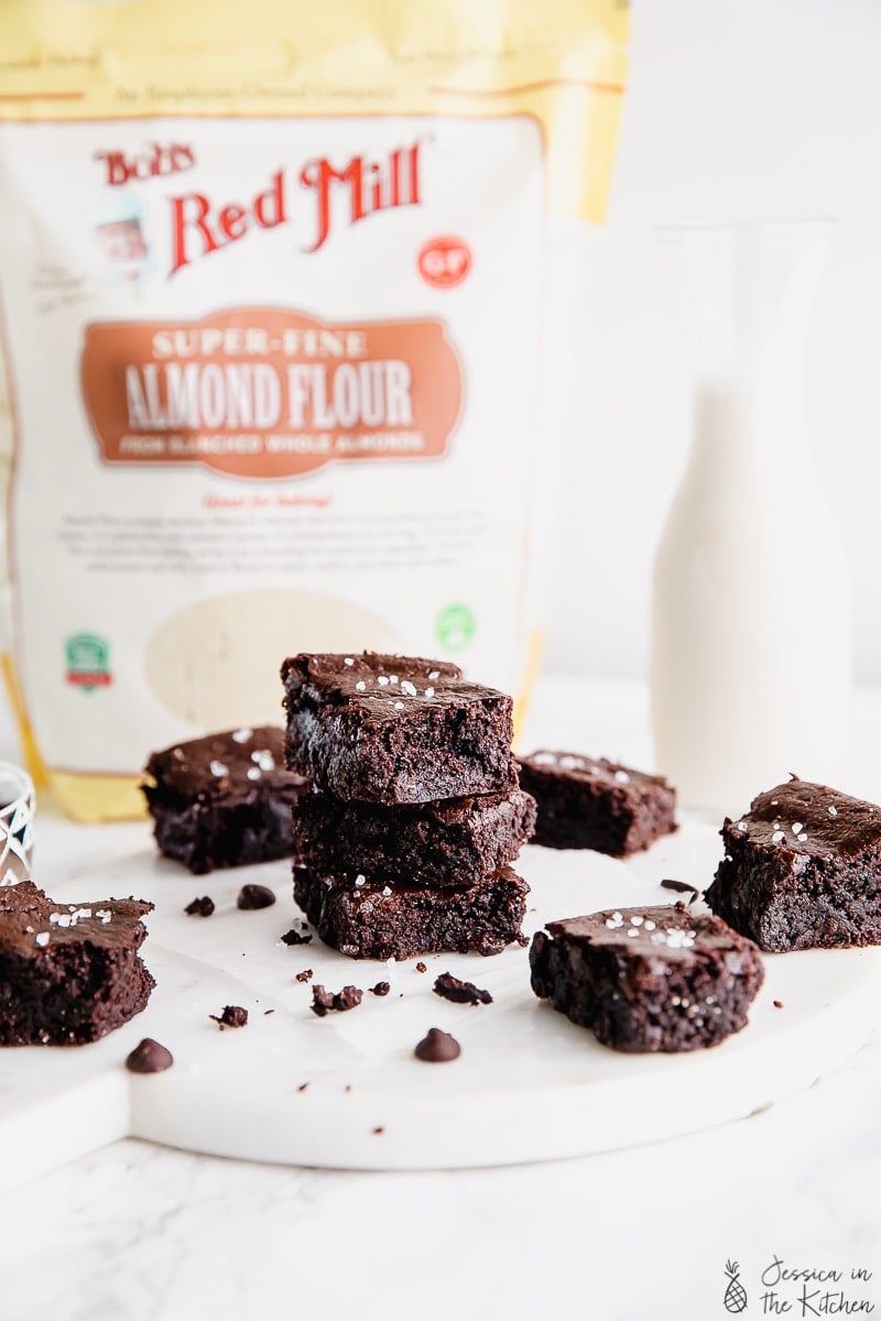 Vegan Almond Flour Brownies impilati uno sull'altro circondati da altri brownies di fronte al sacchetto di farina di mandorle.