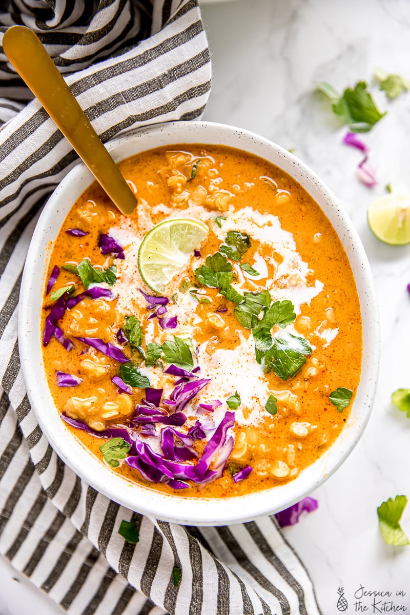 Top down view of lentil soup with lime wedges on the side, topped with cabbage, herbs, coconut milk, and lime, with a spoon in the bowl. 