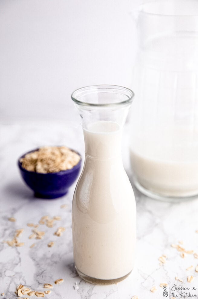 Side shot of homemade oat milk in a milk jar with a container of oats in the background and a larger jug of oat milk in the background.