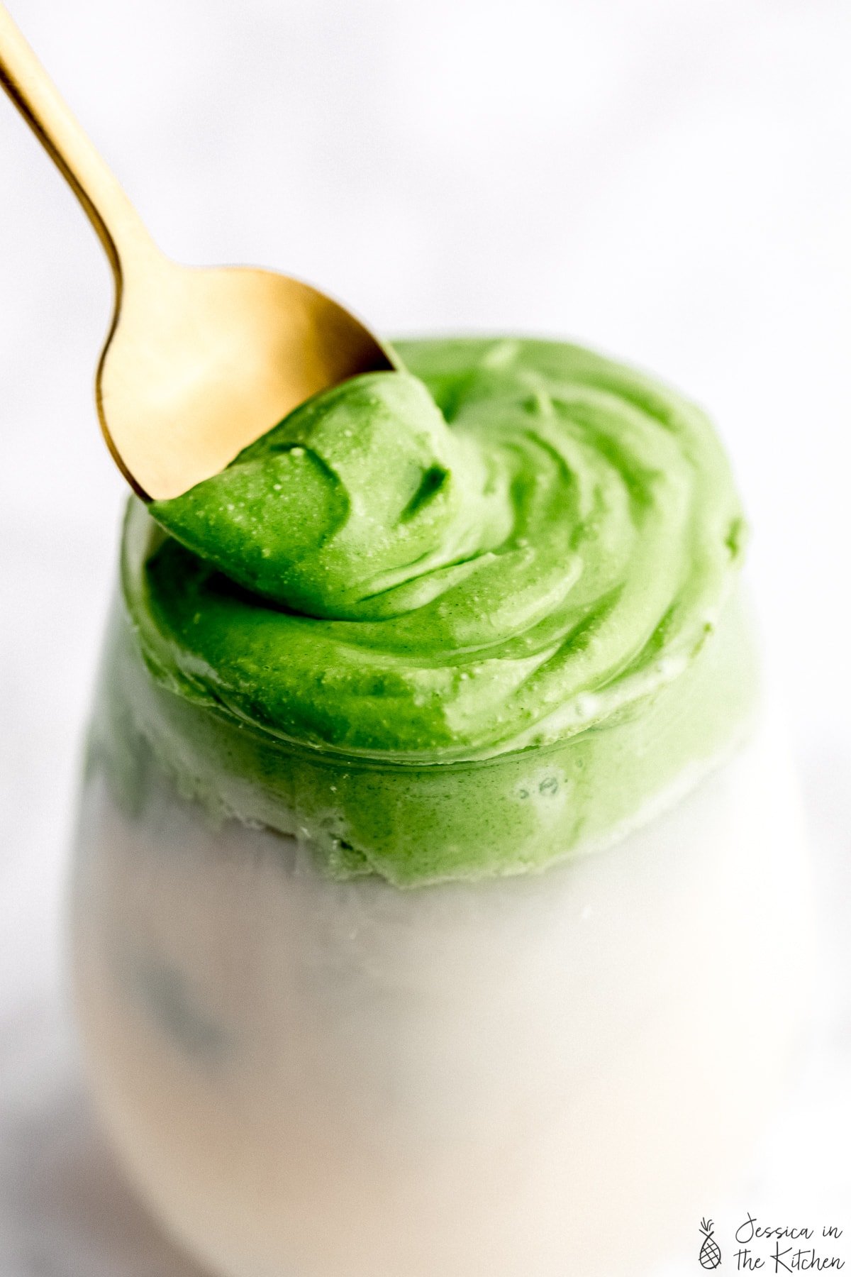 A close up shot of whipped matcha in a glass with milk with a gold spoon slightly pulling the whipped matcha froth.