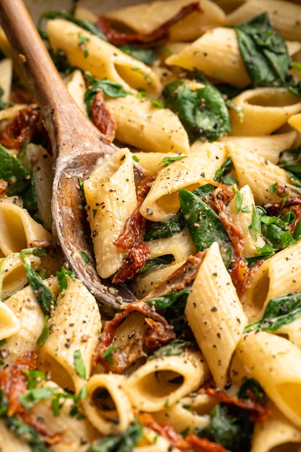 Wooden spoon stirring hummus pasta with sun-dried tomatoes and spinach.