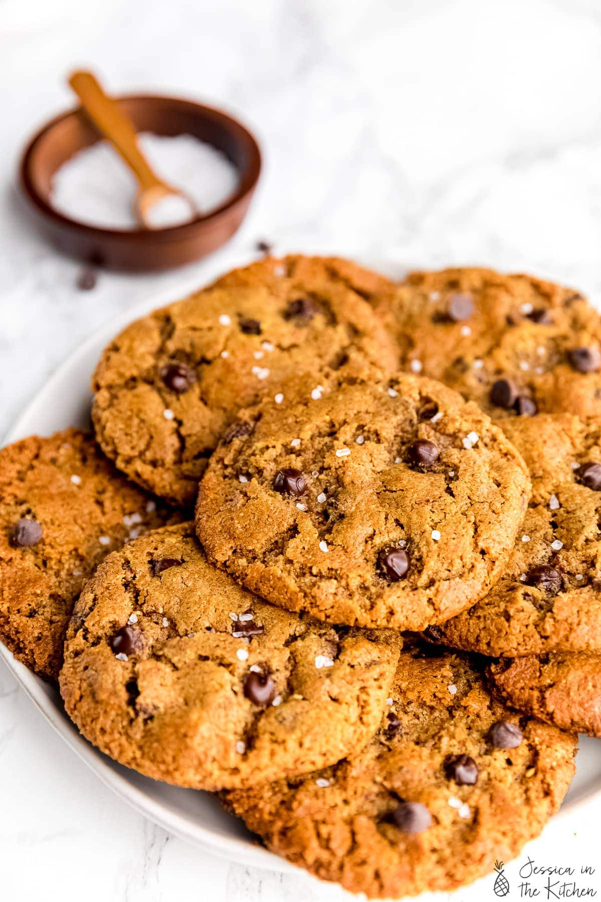Close up of vegan chocolate chip cookies piled on a plate with a bowl of sea salt in the background.