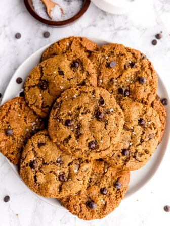 Five ingredient chocolate chip cookies stacked on each other on a plate in front of a jar of salt