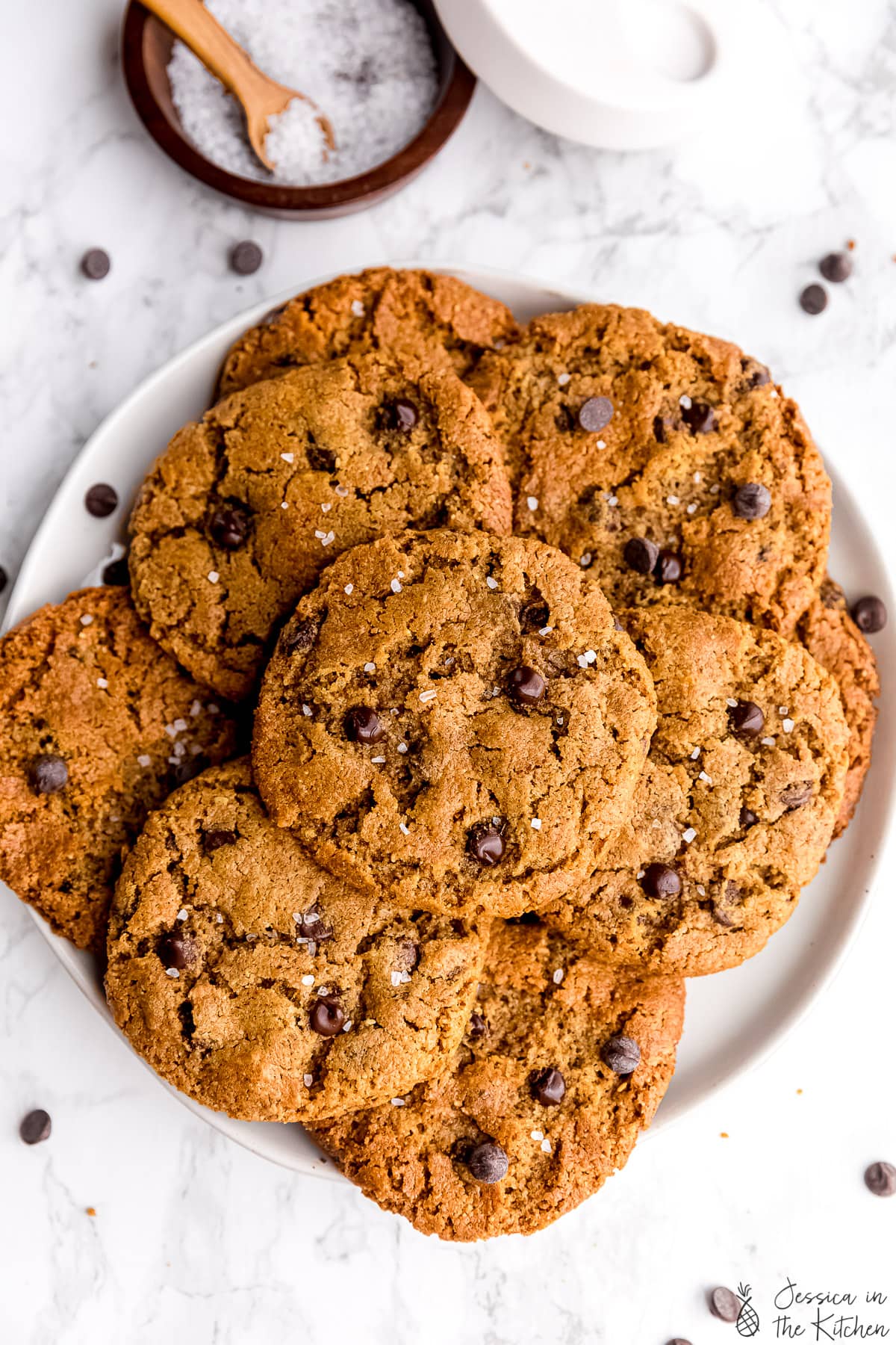 Five ingredient chocolate chip cookies stacked on each other on a plate in front of a jar of salt