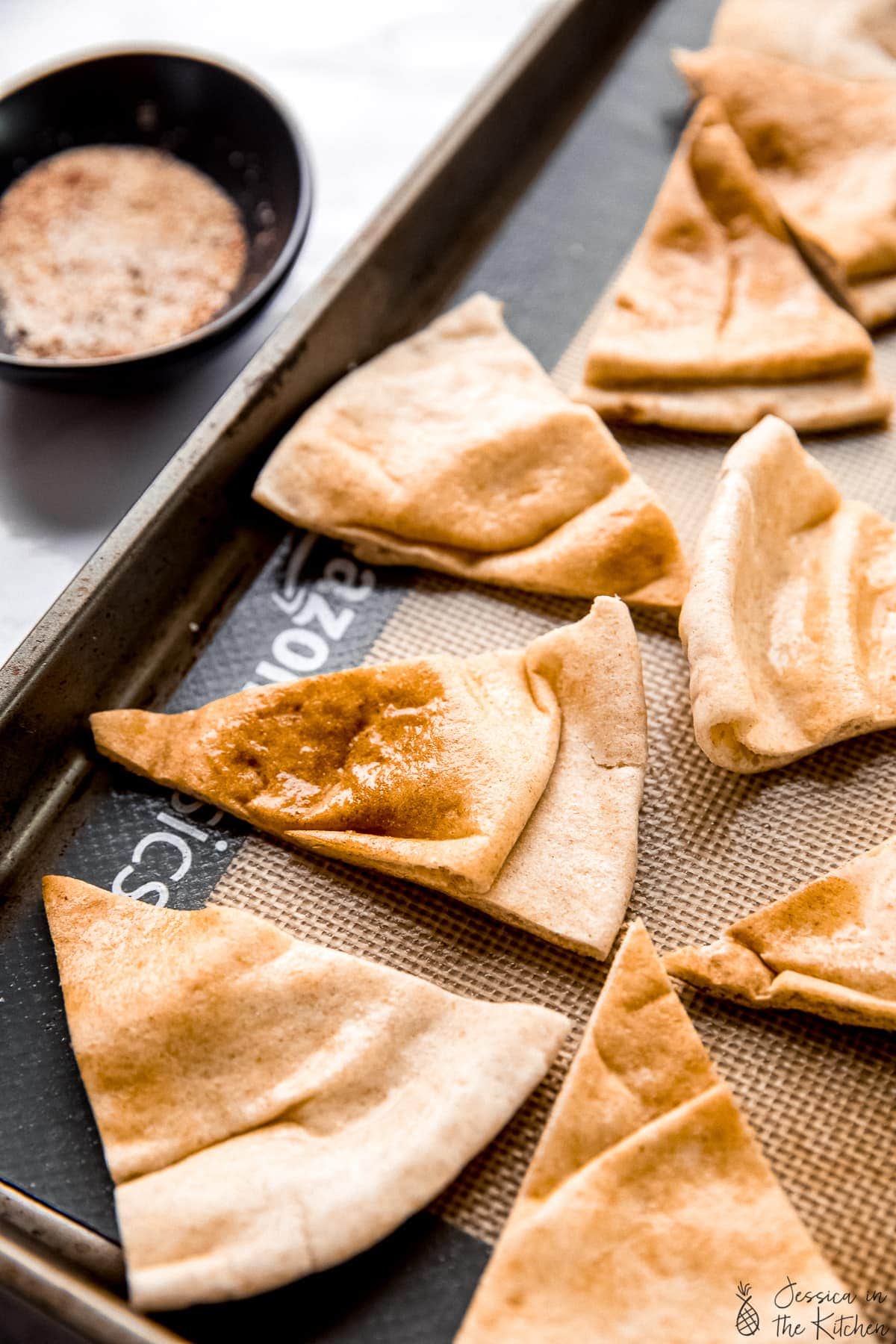 Sliced pita chips brushed with oil on a baking sheet with seasoning mix in a black bowl beside it on a counter top.