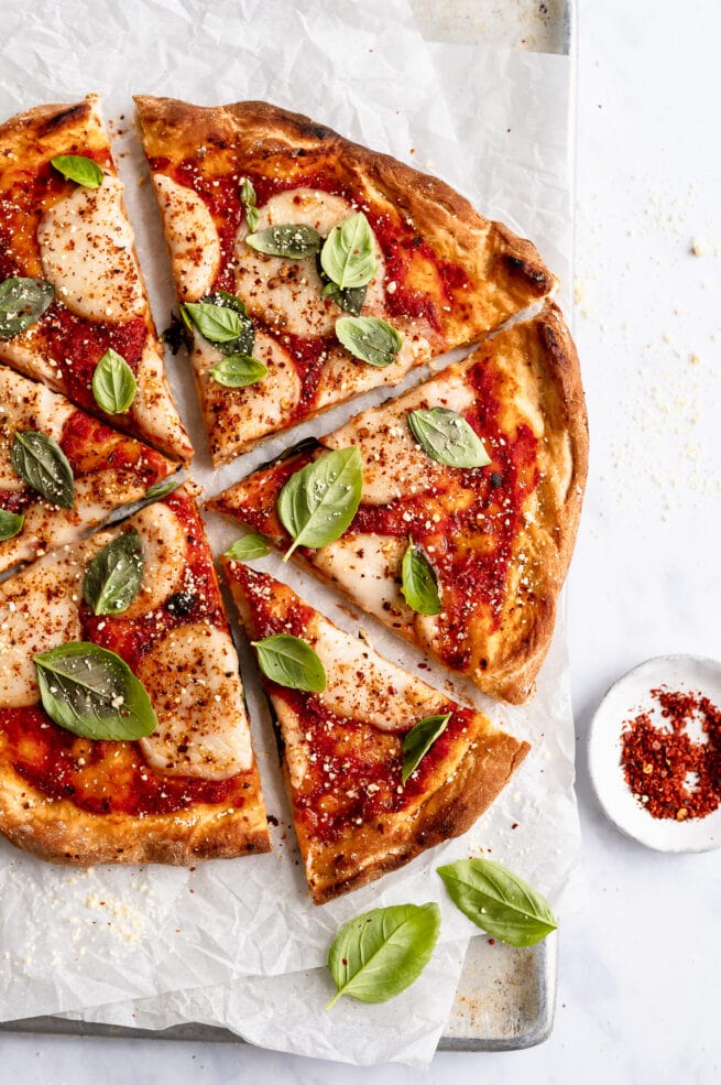 overhead closeup shot of vegan pizza sliced with one slice slightly pulled out. Scattered basil around, and on parchment paper on a baking sheet