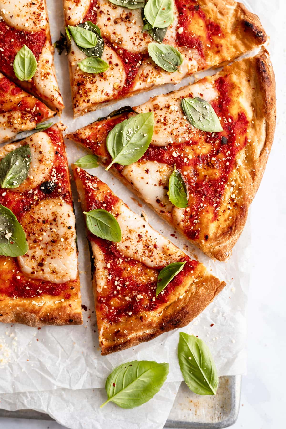 overhead closeup shot of vegan pizza sliced with one slice slightly pulled out. Scattered basil around, and on parchment paper on a baking sheet