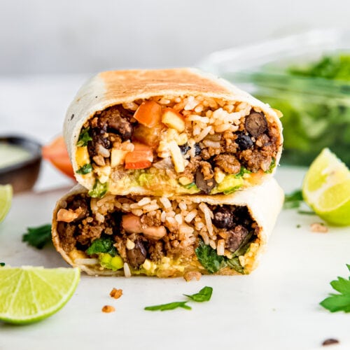 stacked Vegan burrito on a white cutting board surrounded by lime, parsley and ingredients