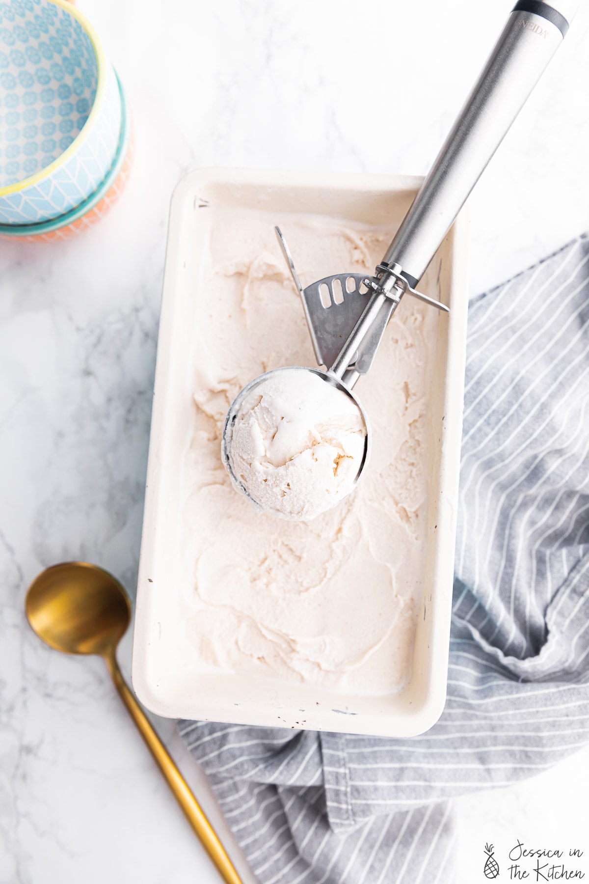 Overhead photo of ice cream in an ice cream scoop in a larger container of ice cream with surrounding bowls, kitchen towel and spoon.