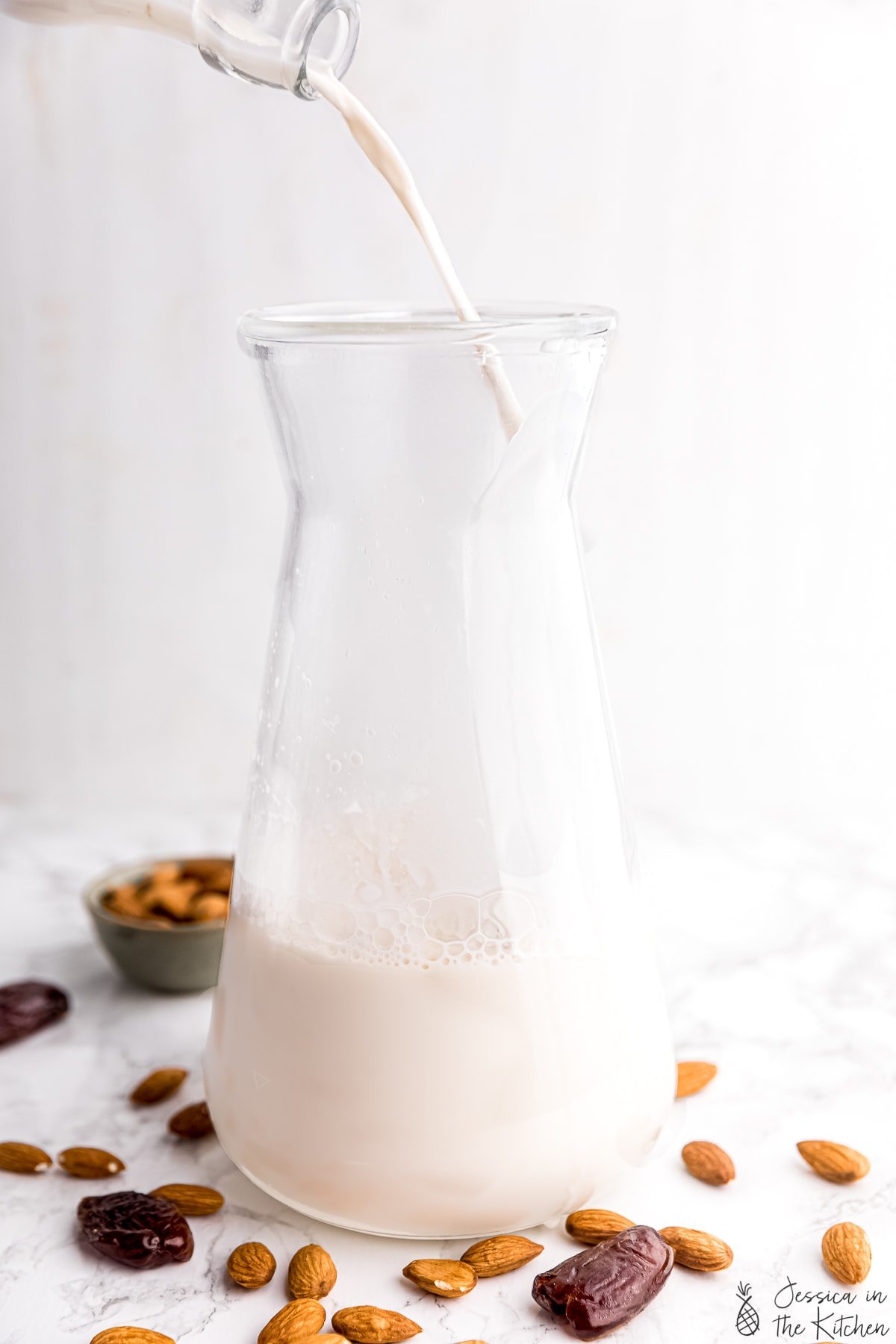 Photo of almond milk being poured into a jar with raw almonds and dates surrounding it.