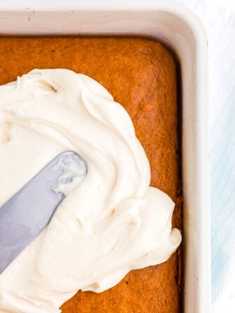 Cream cheese frosting spread across an orange pumpkin cake with a silver offset spatula.