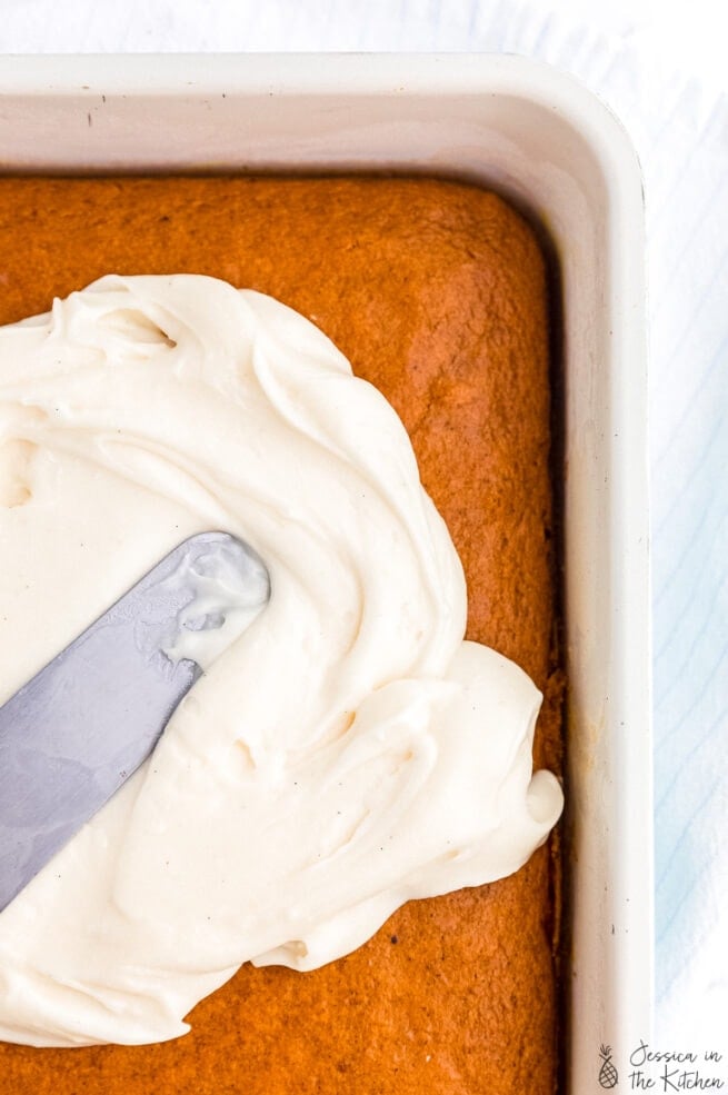 Cream cheese frosting spread across an orange pumpkin cake with a silver offset spatula.