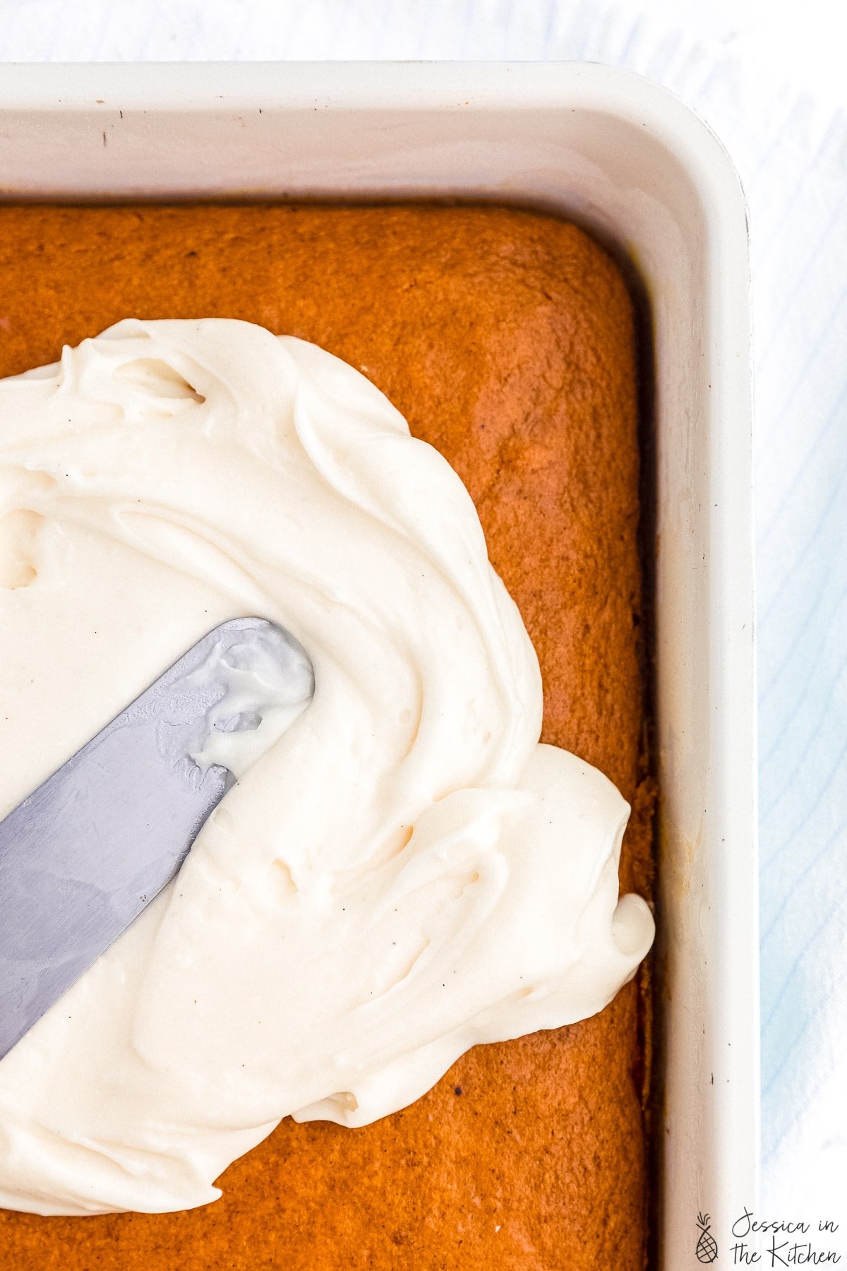 Cream cheese frosting being spread across an orange pumpkin cake with a silver offset spatula.