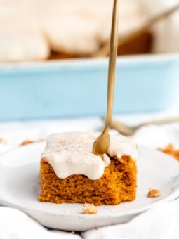 A fork being stuck into a pumpkin cake on a plate with a bite missing in front of a blue container filled with slices of cake.