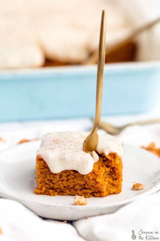 A fork being stuck into a pumpkin cake on a plate with a bite missing in front of a blue container filled with slices of cake.