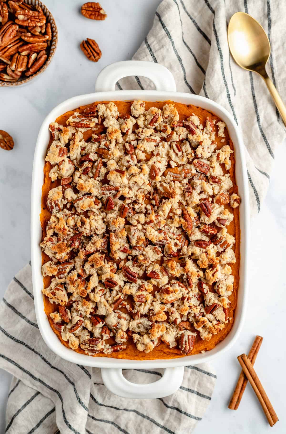 Sweet potato casserole on a striped kitchen towel surround by ingredients and a gold spoon.