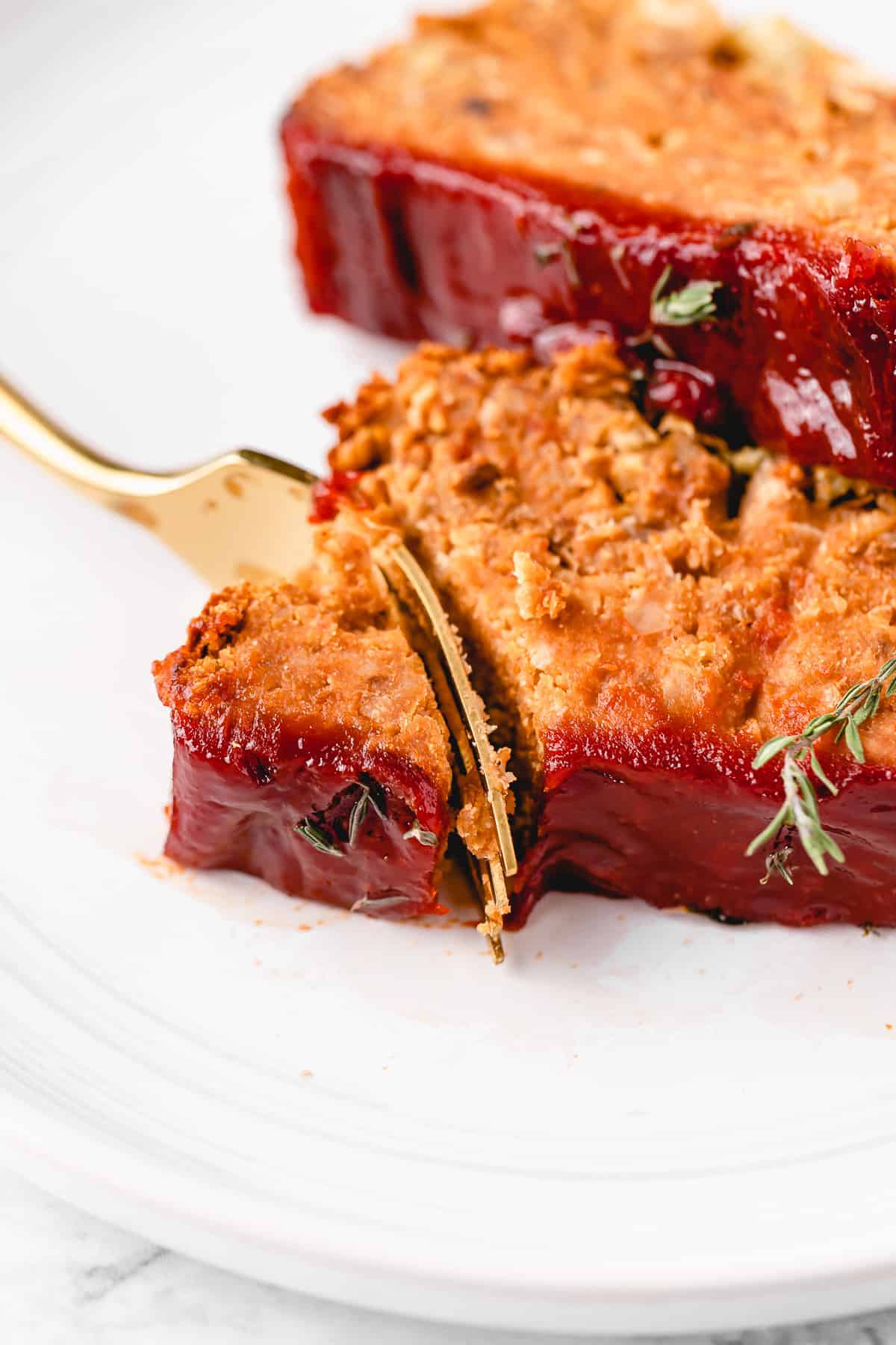 Close up of a fork cutting into the corner of a slice of vegan meatloaf, with a second slice in the background.