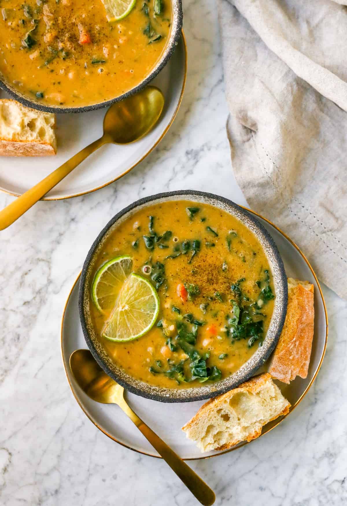 a bowl of white bean soup on a plate with a gold spoon inside, bread, lime and kale