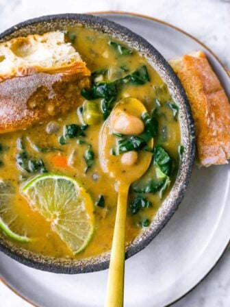 a bowl of white bean soup with a gold spoon inside, bread, lime and kale