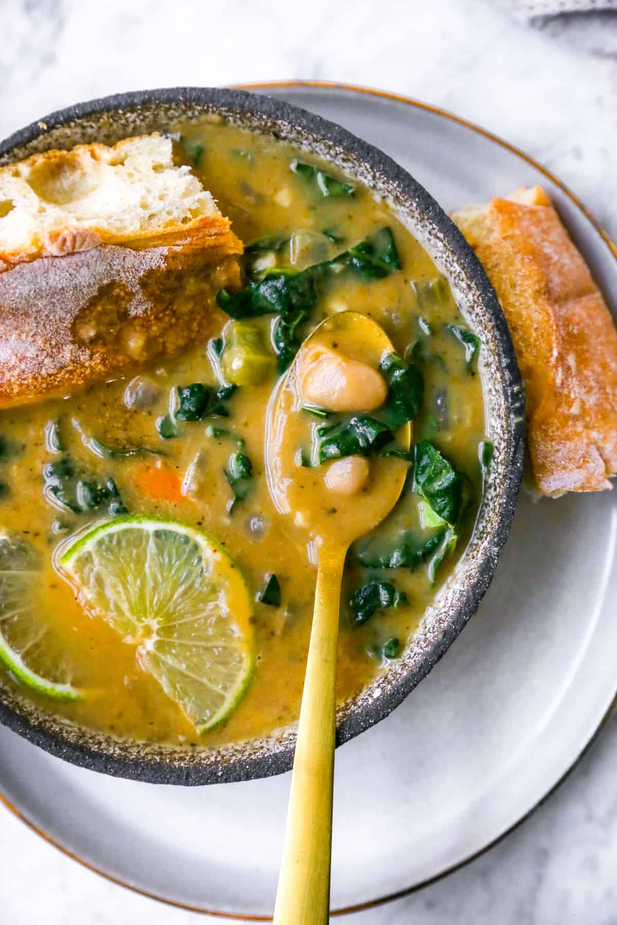 a bowl of white bean soup with a gold spoon inside, bread, lime and kale