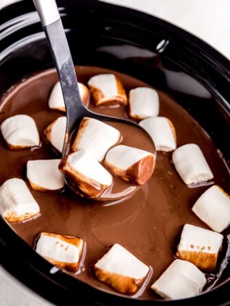a ladle in a slow cooker filled with hot cocoa and vegan marshmallows with some cocoa and three marshmallows on ladle