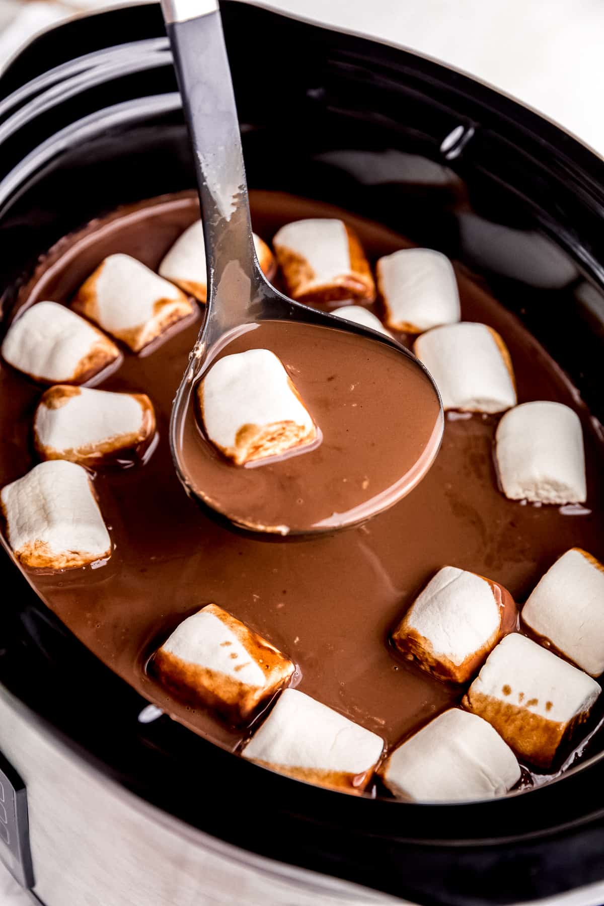 a ladle in a slow cooker filled with hot cocoa and vegan marshmallows with some cocoa and one marshmallow on ladle