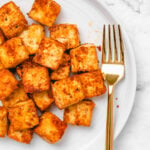 Close-up shot of crispy seasoned tofu on a round white plate with a gold fork