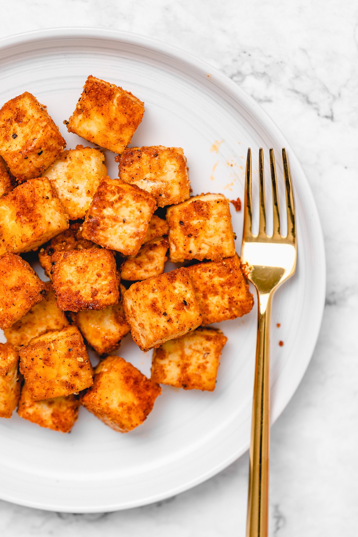 Close-up shot of crispy seasoned tofu on a round white plate with a gold fork