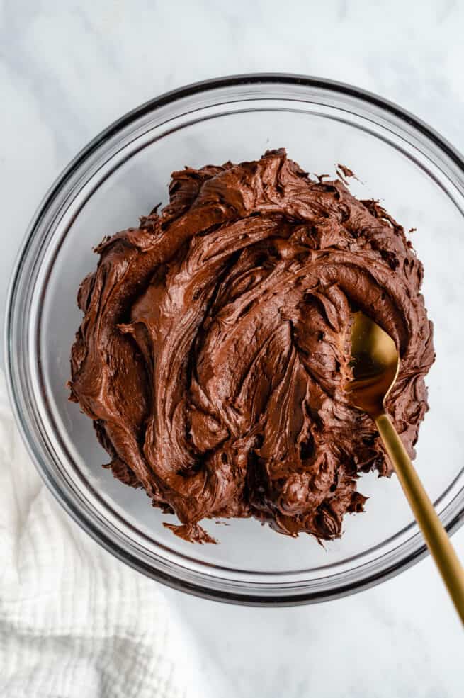 Glass mixing bowl of rich chocolate frosting with a gold spoon