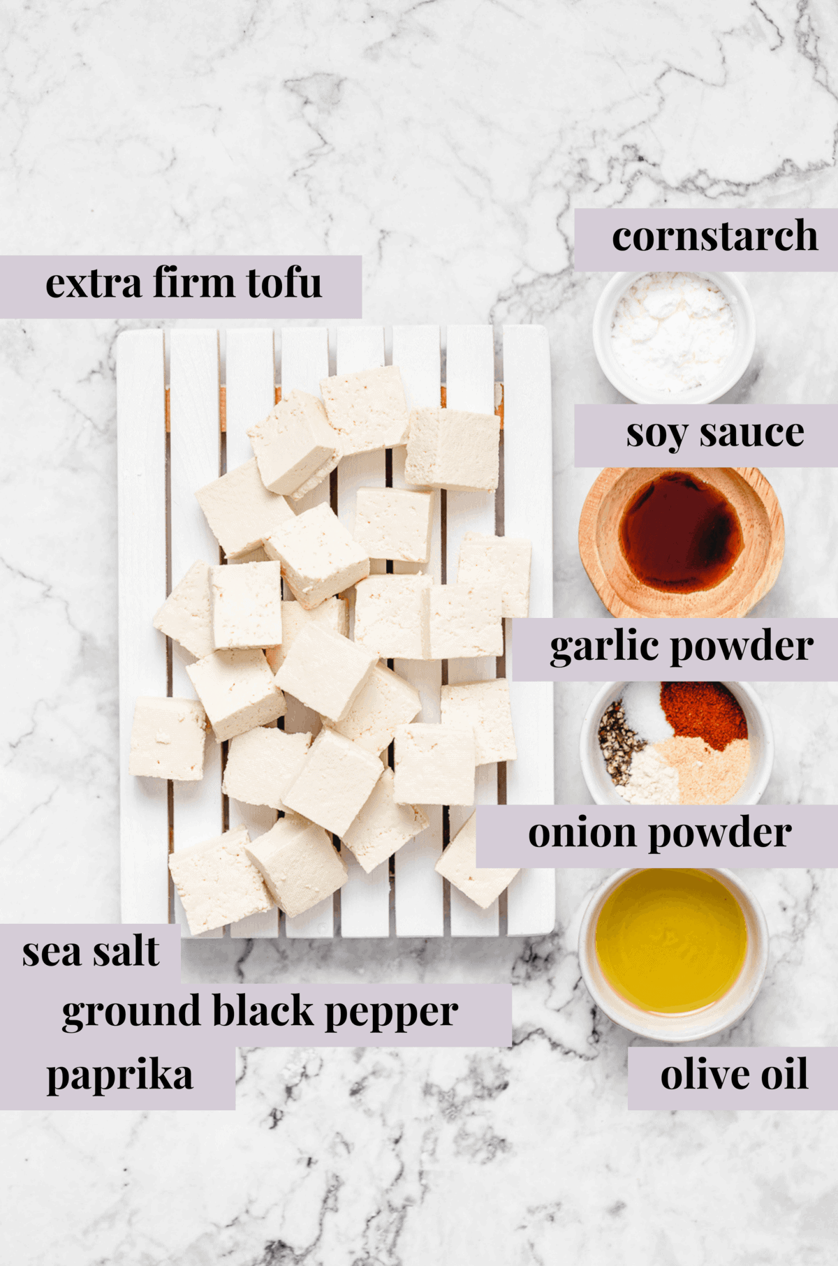 Cubed tofu on a white cutting board surrounded by cornstarch, soy sauce, seasonings, and olive oil