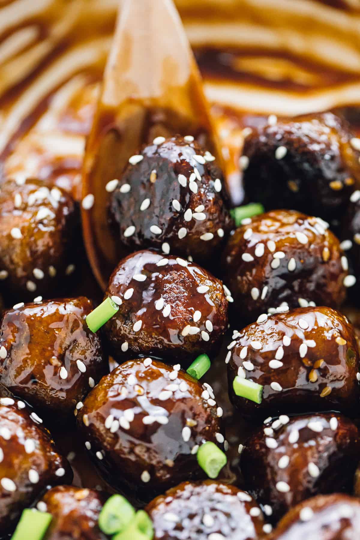 A wooden spoon stirring a pan of saucy vegan meatballs garnished with sesame seeds and scallions