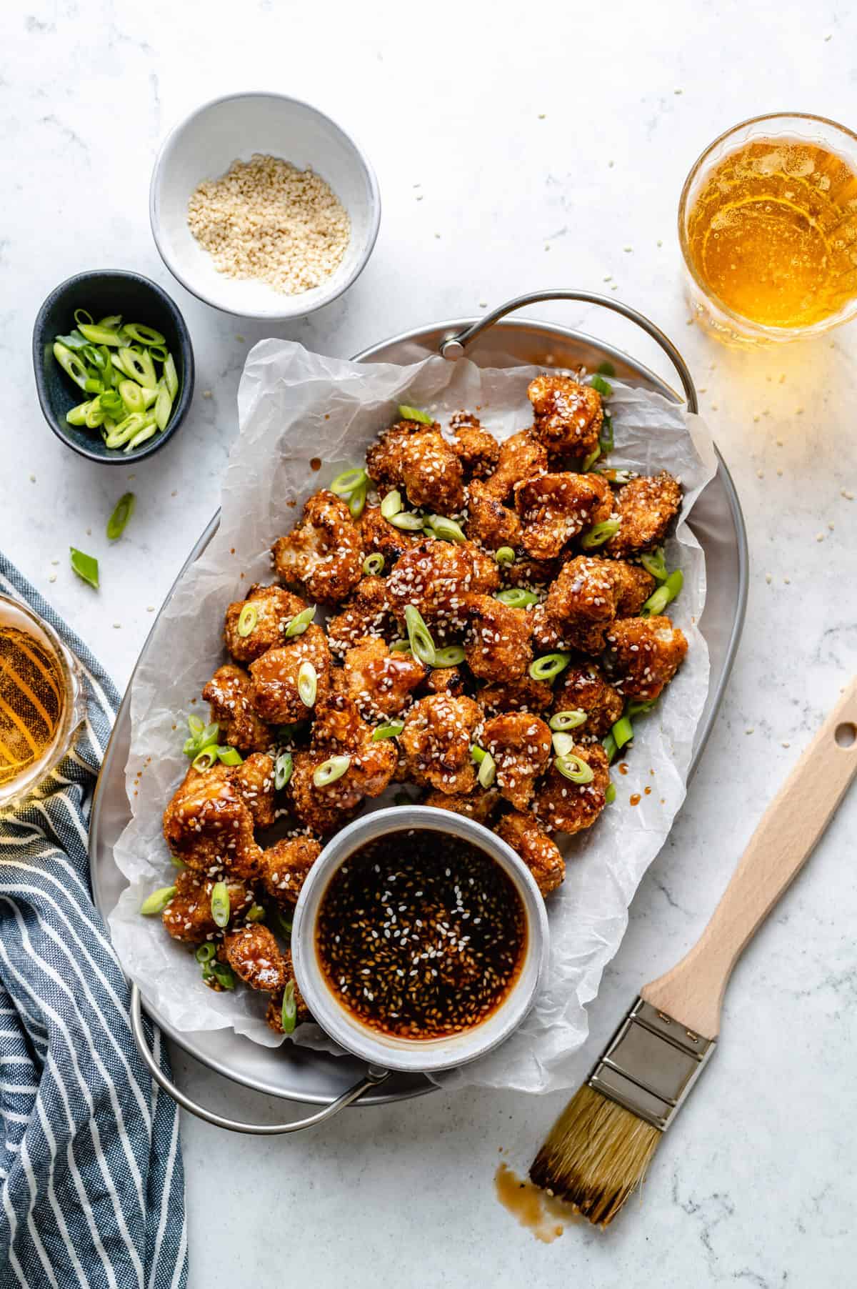plate of cauliflower wings in a tray with sauce in a small bowl beside it surrounding by toppings and a brush to brush sauce on wings