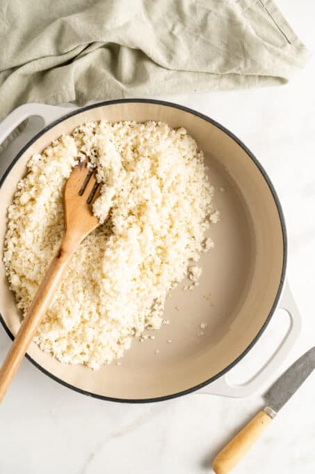 Overhead view of uncooked cauliflower rice in white cast iron skillet with wooden spatula