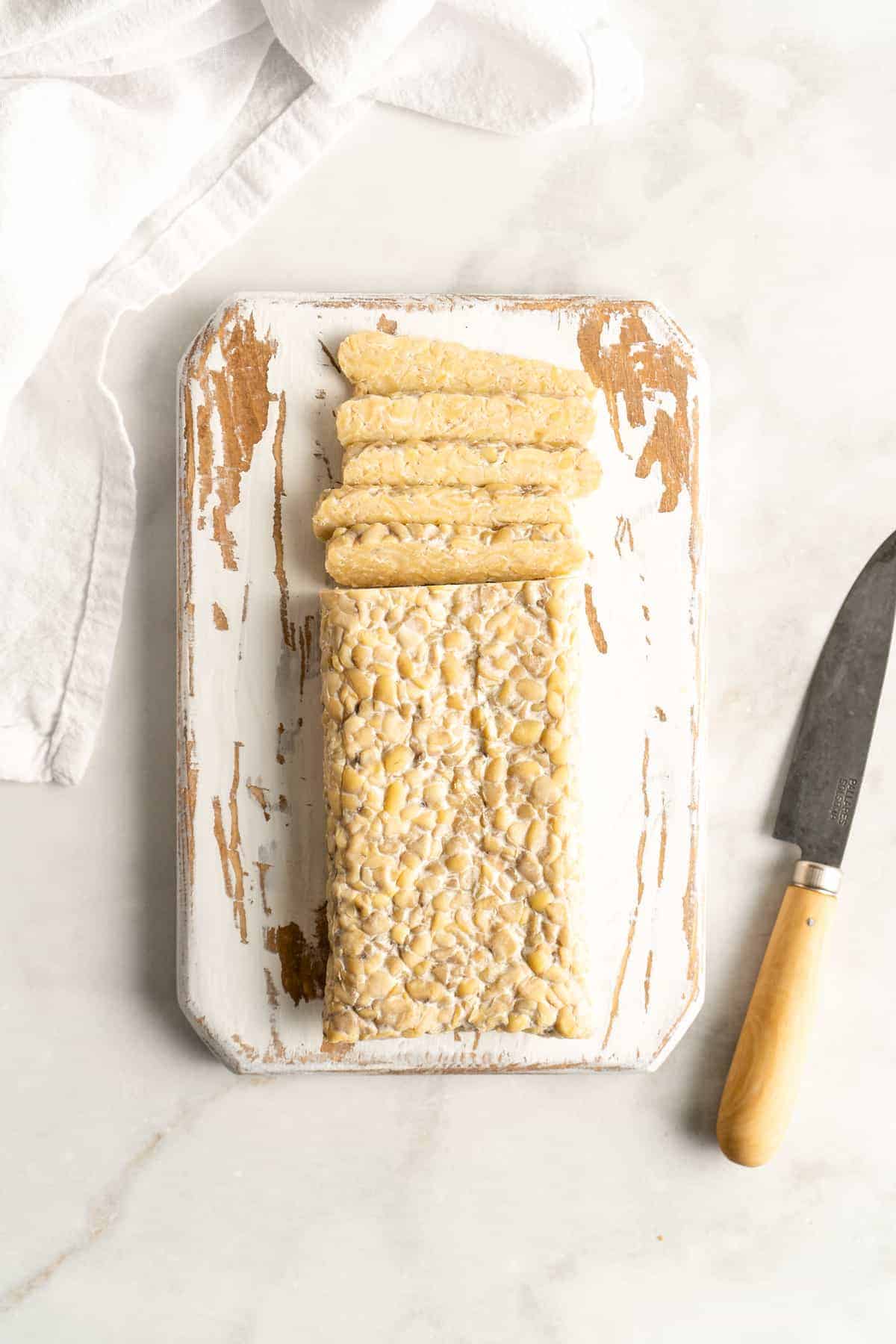 Overhead shot of tempeh being sliced on rustic white painted cutting board