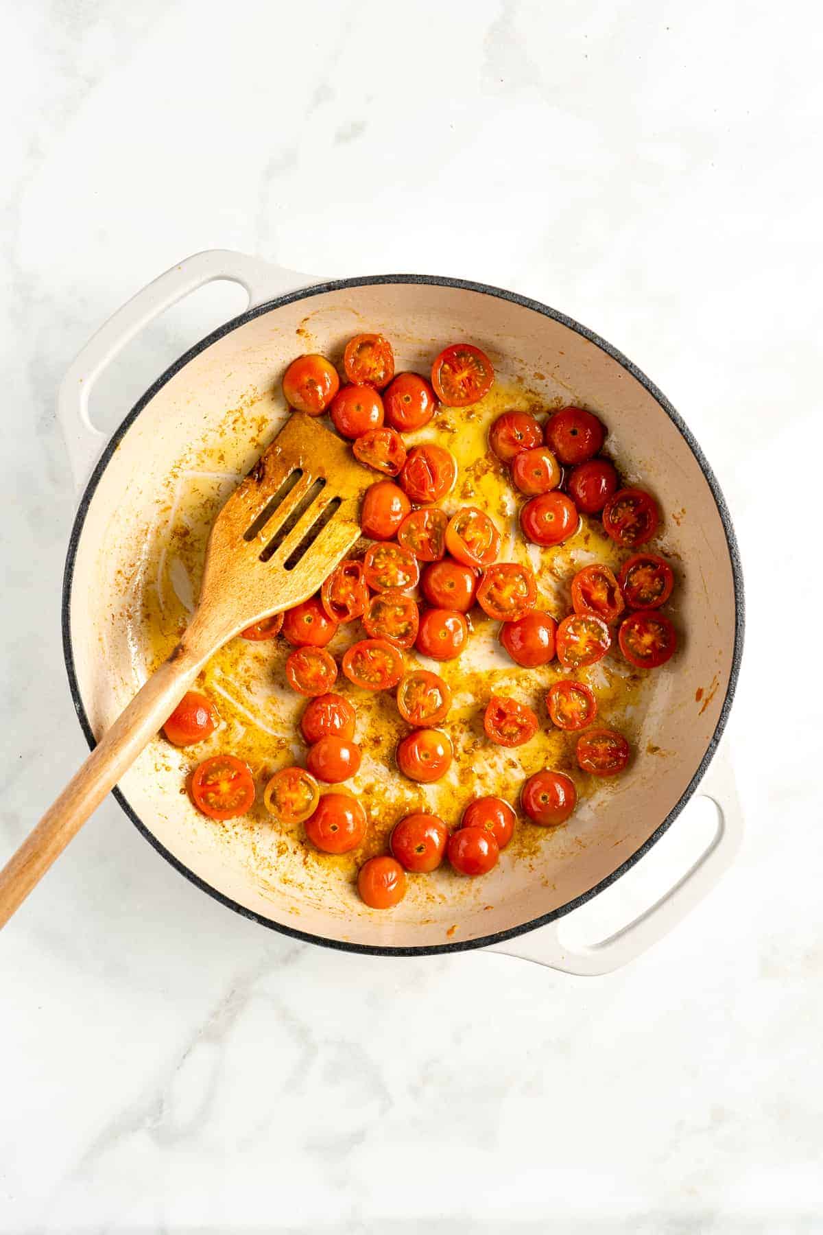 Overhead view of cooked cherry tomatoes and wooden spatula in double handled white cooking pot