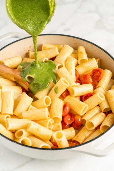 Pesto sauce being poured onto cooked rigatone pasta and cherry tomatoes inside of a double handled white cooking pot