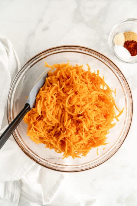 Glass bowl filled with shredded sweet potatoes, with small bowl of seasonings in background