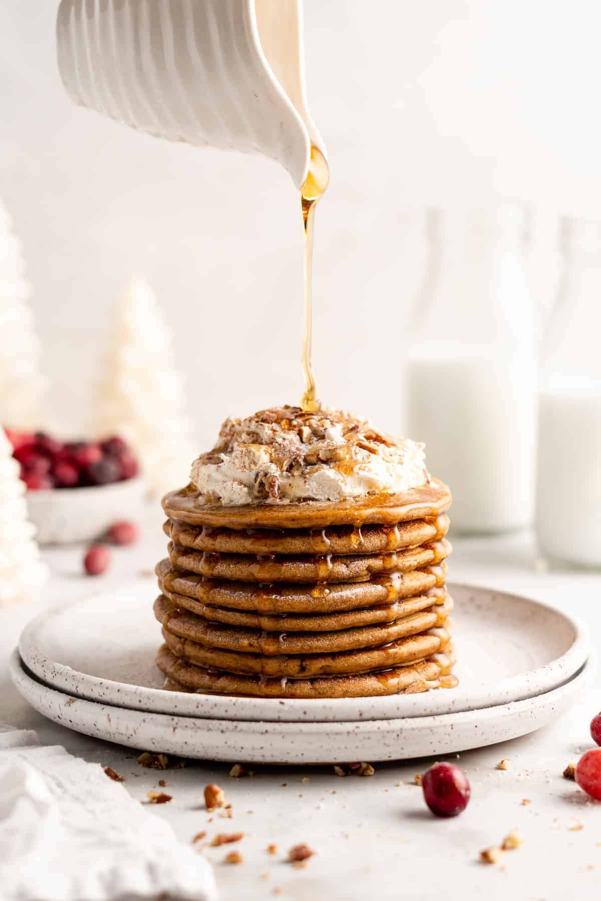 Pouring maple syrup over stack of pancakes topped with coconut whipped cream and nuts