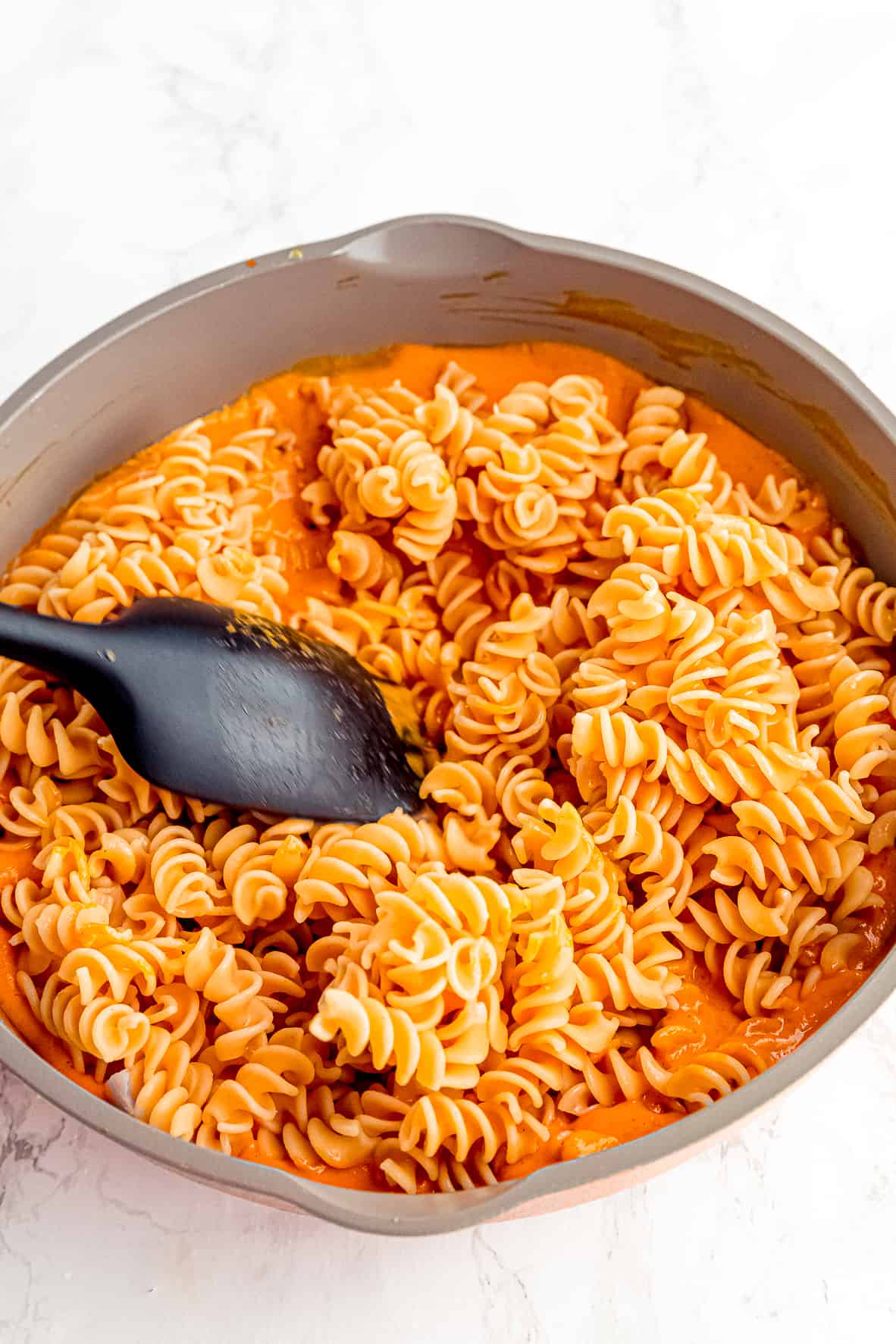 A spatula stirring cooked pasta into roasted red pepper pasta sauce in a skillet.