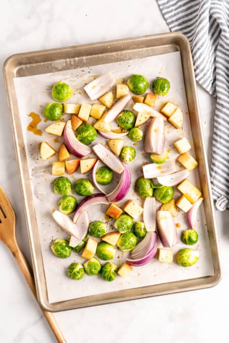 Overhead view of apples, Brussels sprouts, and red onions on a parchment lined sheet pan.