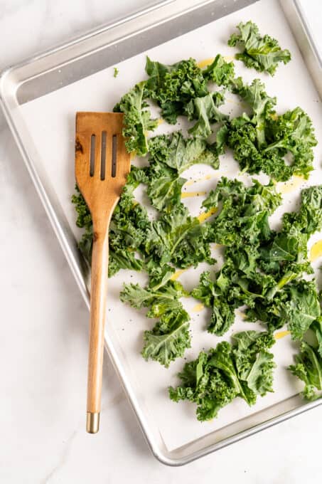 Overhead view of kale leaves on a parchment-lined sheet pan with a wooden spatula.