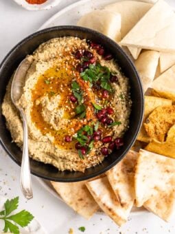 Overhead view of baba ghanoush in black bowl with spoon, surrounded by pitas