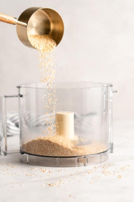 Measuring cup of sesame seeds being poured into food processor bowl