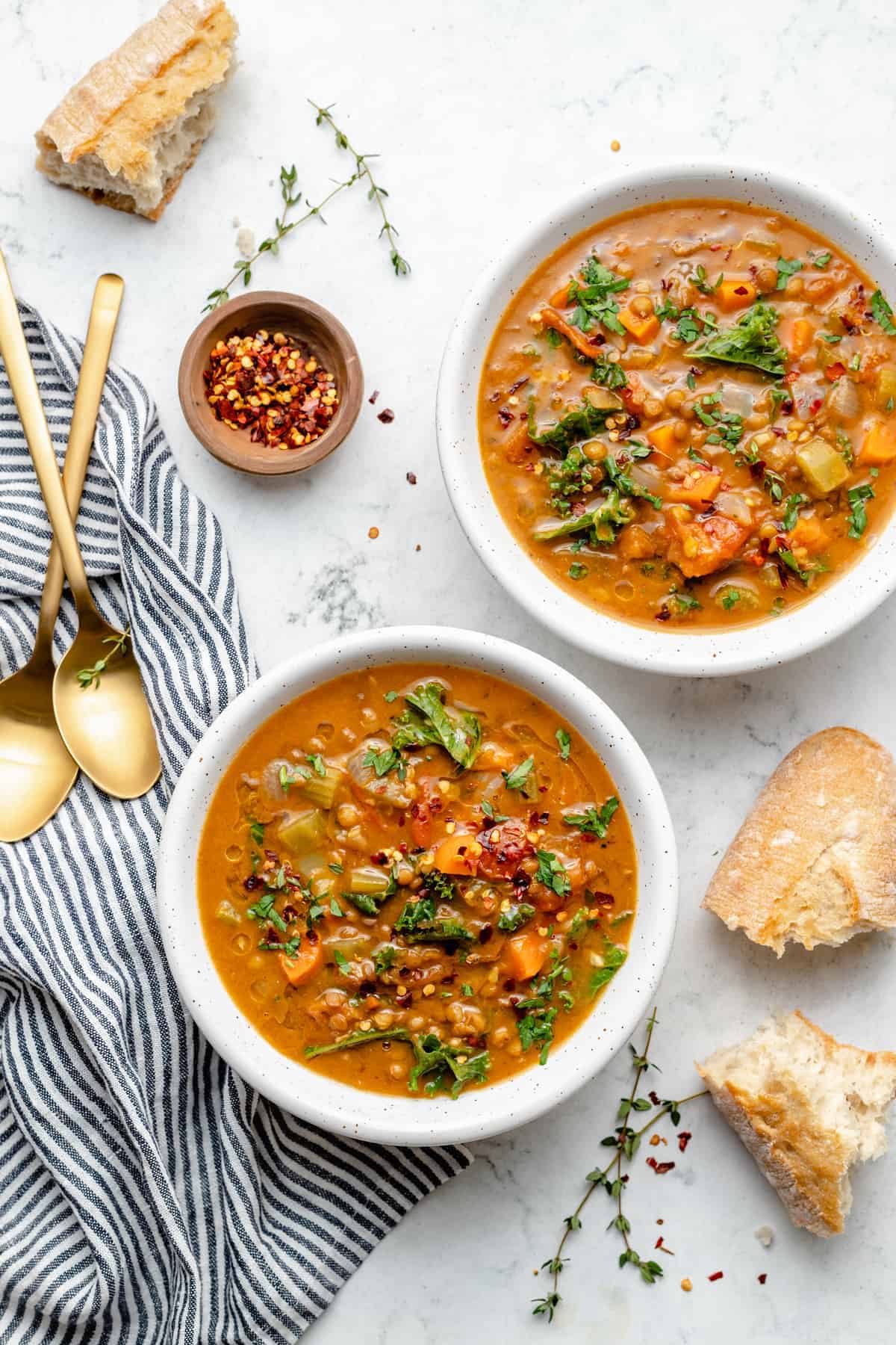 Overhead view of two bowls of lentil soup on marble board with baguette pieces and garnishes