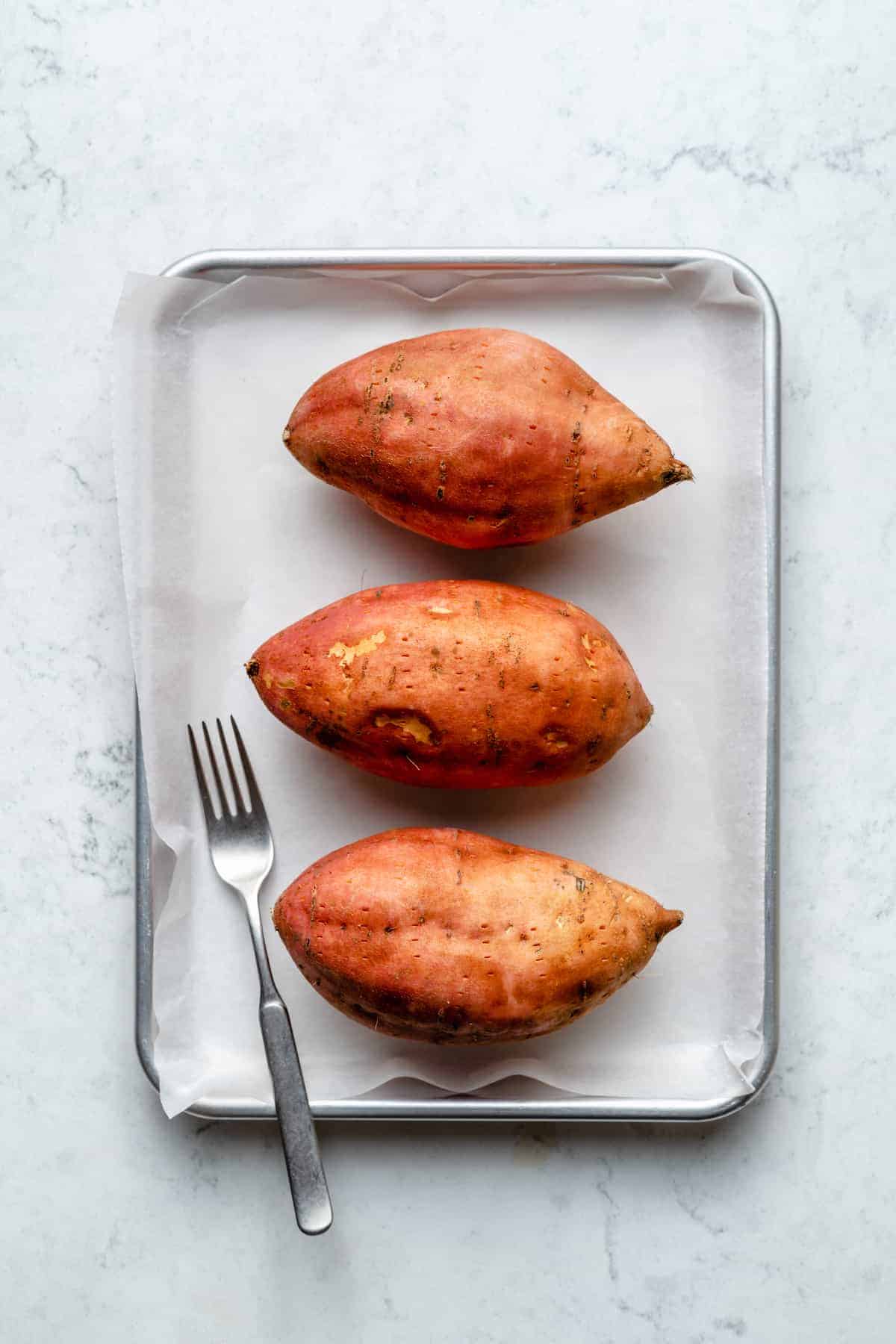 Overhead view of 3 sweet potatoes on baking sheet with fork