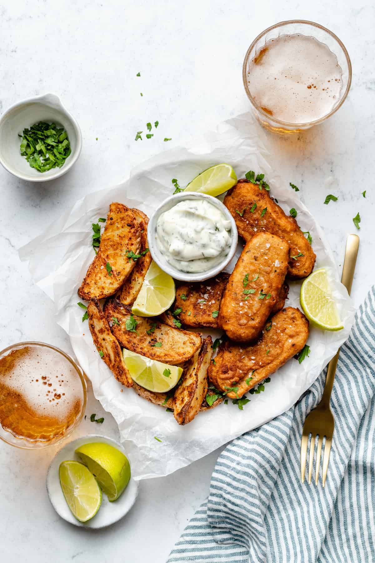 Overhead view of vegan fish sticks on plate surrounded by lime wedges, glasses of beer, and chopped parsley